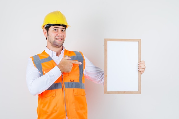 Homem construtor apontando o dedo para o quadro branco na camisa, uniforme e olhando alegre, vista frontal.