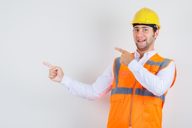 Homem construtor apontando os dedos para o lado na camisa, uniforme e parecendo alegre, vista frontal.