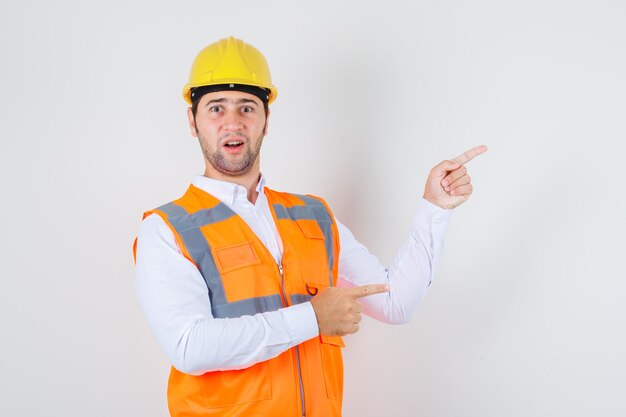 Homem construtor apontando os dedos para o lado na camisa, uniforme e parecendo espantado, vista frontal.