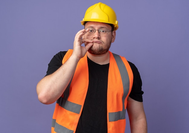 Homem construtor com colete de construção e capacete de segurança fazendo gesto de silêncio com os dedos, como fechar a boca com um zíper de pé sobre um fundo azul