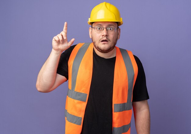 Homem construtor com colete de construção e capacete de segurança olhando para a câmera surpreso e preocupado, mostrando o dedo indicador em pé sobre o azul