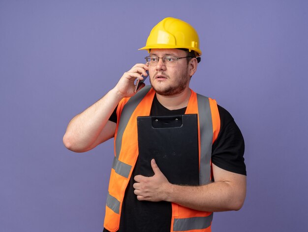 Homem construtor em colete de construção e capacete de segurança segurando a prancheta, olhando para o lado com uma cara séria enquanto fala ao telefone móvel em pé sobre um fundo azul