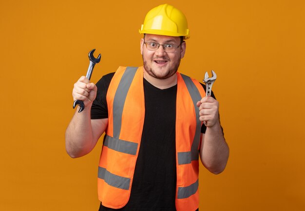 Homem construtor em colete de construção e capacete de segurança segurando palavras olhando para a câmera feliz e surpreso em pé sobre um fundo laranja