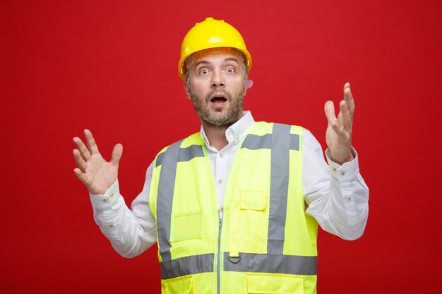 Homem construtor em uniforme de construção e capacete de segurança olhando para a câmera preocupado e confuso levantando os braços sem resposta em pé sobre fundo vermelho