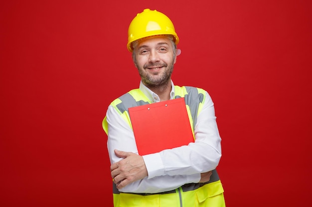 Foto grátis homem construtor em uniforme de construção e capacete de segurança segurando a área de transferência olhando para a câmera feliz e positiva sorrindo em pé sobre fundo vermelho