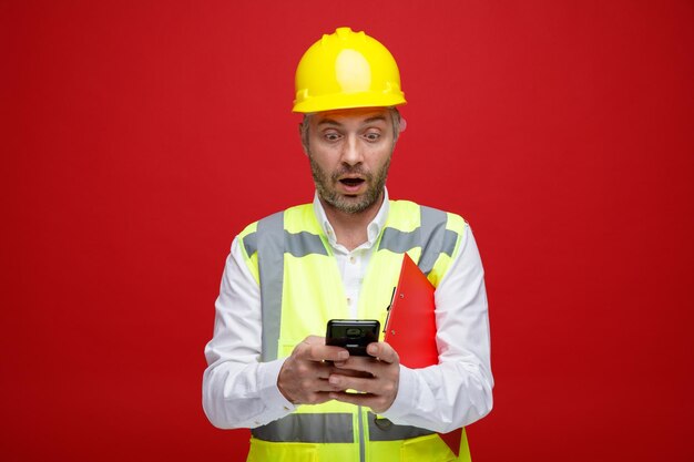 Homem construtor em uniforme de construção e capacete de segurança segurando mensagem de texto da área de transferência usando smartphone olhando espantado e surpreso em pé sobre fundo vermelho