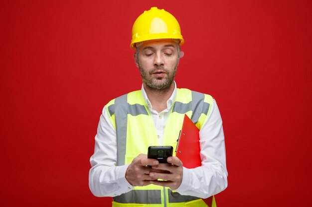 Homem construtor em uniforme de construção e capacete de segurança segurando mensagem de texto da área de transferência usando smartphone parecendo confiante em pé sobre fundo vermelho
