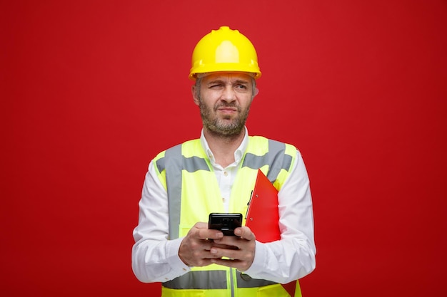 Homem construtor em uniforme de construção e capacete de segurança segurando mensagem de texto da área de transferência usando smartphone parecendo descontente fazendo boca torta em pé sobre fundo vermelho