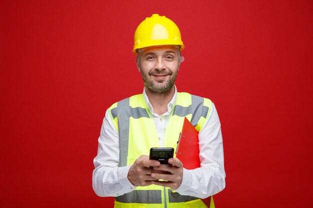 Homem construtor em uniforme de construção e capacete de segurança segurando mensagem de texto da área de transferência usando smartphone sorrindo feliz e positivo olhando para a câmera em pé sobre fundo vermelho