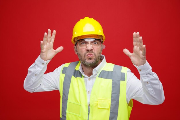 Homem construtor em uniforme de construção e capacete de segurança usando óculos de segurança irritado e frustrado levantando os braços enlouquecendo em pé sobre fundo vermelho