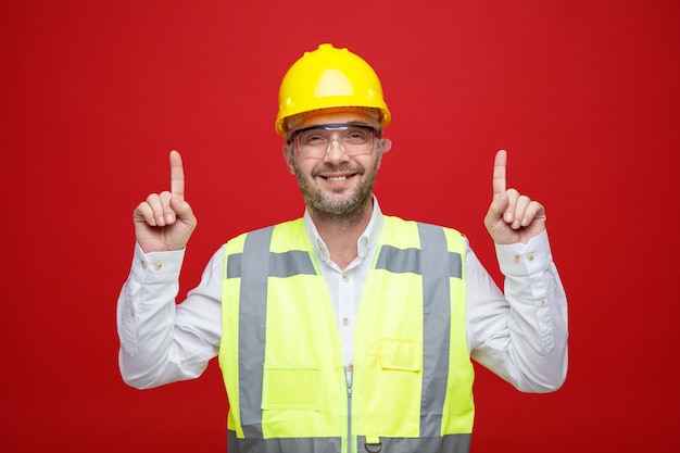 Homem construtor em uniforme de construção e capacete de segurança usando óculos de segurança olhando para a câmera feliz e alegre apontando com os dedos indicadores para cima em pé sobre fundo rosa