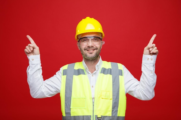 Foto grátis homem construtor em uniforme de construção e capacete de segurança usando óculos de segurança, olhando para a câmera sorrindo feliz e positivo apontando com os dedos indicadores em pé sobre fundo rosa