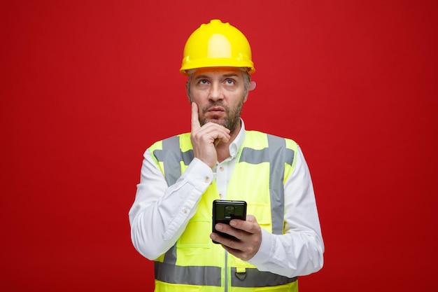 Homem construtor em uniforme de construção e mensagem de texto de capacete de segurança usando smartphone olhando intrigado em pé sobre fundo vermelho