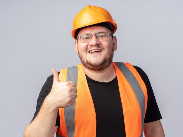 Homem construtor feliz e positivo em colete de construção e capacete de segurança olhando para a câmera sorrindo alegremente mostrando os polegares para cima em pé sobre o branco