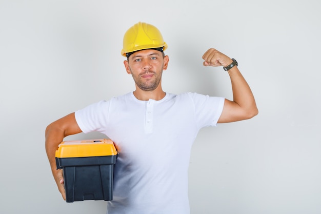 Homem Construtor segurando a caixa de ferramentas e mostrando os músculos em t-shirt, capacete e olhando confiante, vista frontal.