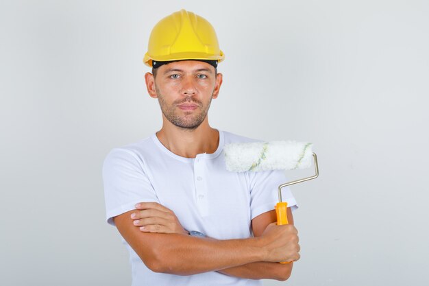 Homem Construtor segurando rolo de pintura com os braços cruzados em camiseta branca, capacete, vista frontal.