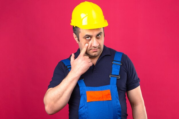 Homem Construtor vestindo uniforme de construção e capacete de segurança, apontando para o olho, observando você gesticular expressão suspeita sobre parede rosa isolada