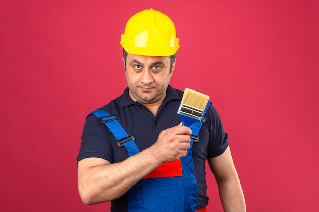 Homem construtor vestindo uniforme de construção e capacete de segurança em pé com pincel com sorriso no rosto sobre parede rosa isolada