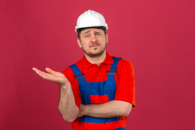 Homem Construtor vestindo uniforme de construção e capacete de segurança, sorrindo para a câmera enquanto se apresenta com a mão em pé sobre a parede rosa isolada