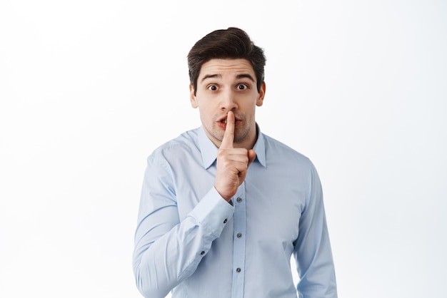 Homem corporativo de camisa pedindo para ficar quieto fofocando no escritório calando segurando o dedo nos lábios e olhando fundo branco animado