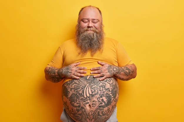 Foto grátis homem corpulento satisfeito mantém as mãos na barriga, sente saciedade após comer um jantar delicioso, fica de pé com os olhos fechados, não liga para a figura, tem desequilíbrio hormonal, isolado na parede amarela.