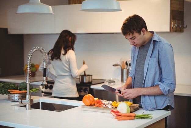 Homem cortando vegetais na cozinha enquanto a mulher cozinhando comida no fundo