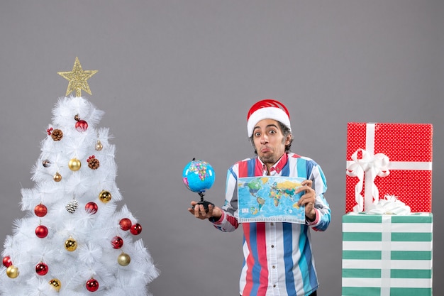 Homem curioso de vista frontal com chapéu de papai noel com mola espiral segurando o mapa-múndi e o globo ao redor da árvore de natal e presentes