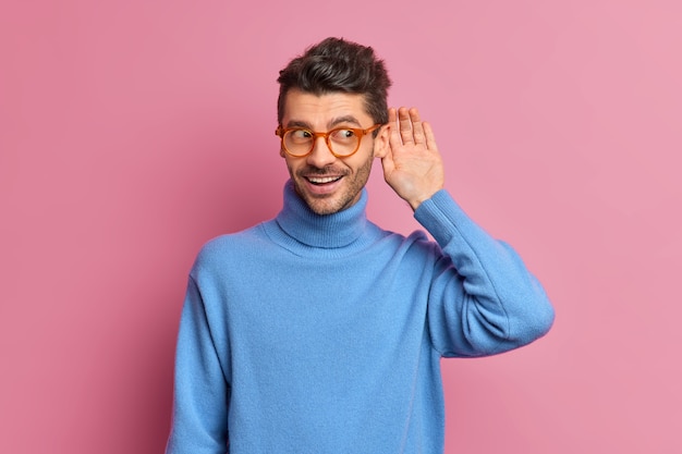 Homem curioso e alegre com a barba por fazer mantém a mão perto do ouvido tentando ouvir uma conversa particular ouve fofocas