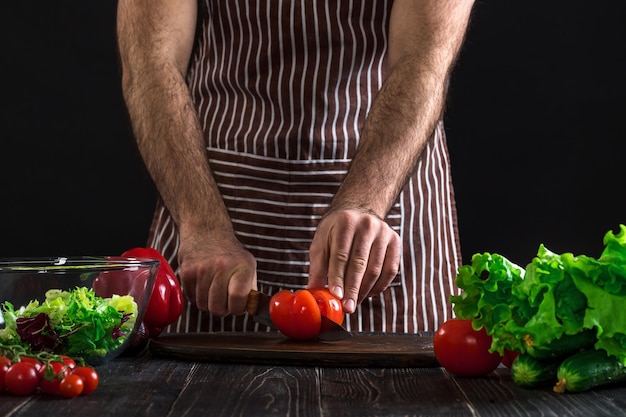 Homem de avental listrado, preparando salada em uma mesa de madeira. as mãos dos homens cortam o tomate para fazer uma salada em fundo preto. conceito de comida saudável