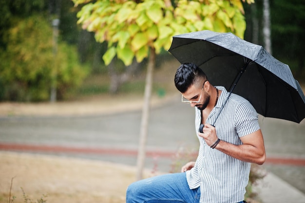 Homem de barba árabe alto na moda usa camisa jeans e óculos de sol com guarda-chuva posou na chuva na praça do parque