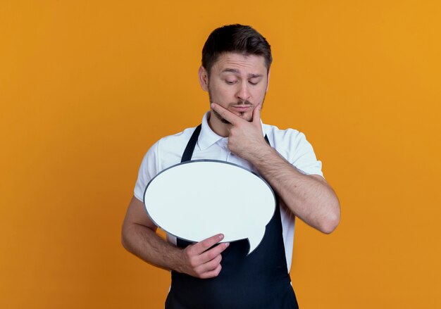 Homem de barbeiro com avental segurando um cartaz de bolha do discurso em branco olhando para ele com a mão no queixo pensando em pé sobre um fundo laranja