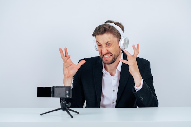 Foto grátis homem de blogueiro com fone de ouvido está mostrando gesto de raiva e mimetismo sentado na frente de sua mini câmera em fundo branco