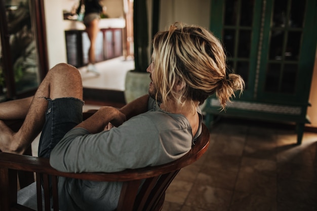 Homem de cabelo comprido, camisa cinza e shorts rasgados senta na cadeira derrubando as pernas na mesa de madeira