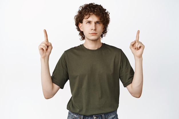 Homem de cabelos encaracolados irritado apontando dedos para cima olhando para o logotipo lendo anúncio com fundo branco de expressão de rosto irritado