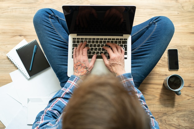 Homem de cabelos escuros com tatuagem rosa na mão digitando no laptop