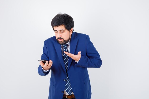 Homem de cabelos escuros em camisa branca, jaqueta azul escura, gravata olhando para o telefone e parecendo indefeso, vista frontal.