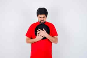 Foto grátis homem de cabelos escuros em uma camiseta vermelha abraçando seu boné e parecendo esperançoso, vista frontal.