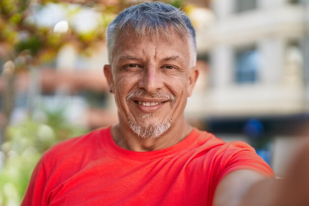 Homem de cabelos grisalhos de meia-idade sorrindo confiante fazendo selfie pela câmera no parque