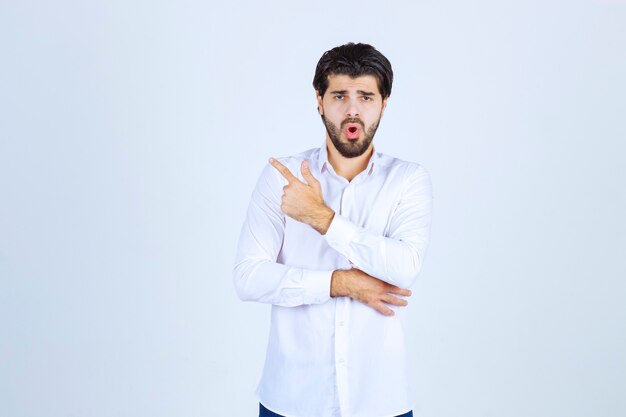 Homem de camisa branca apontando para algum lugar à esquerda com emoções de rosto.