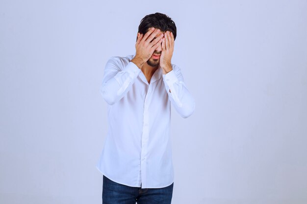 Homem de camisa branca cobrindo o rosto, sente-se triste e tem dor de cabeça.