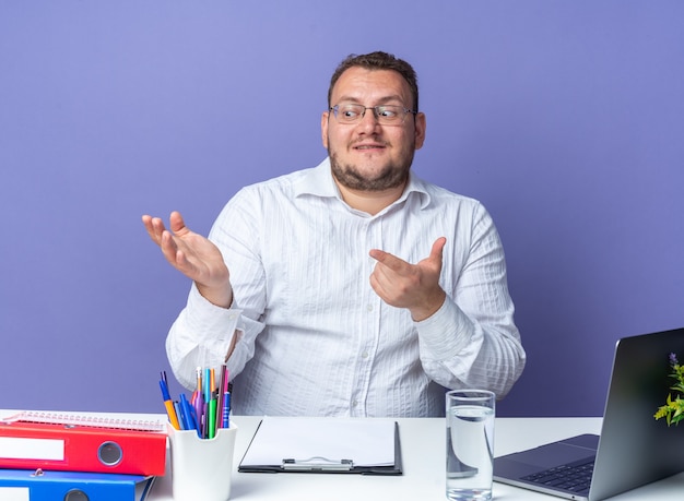 Homem de camisa branca de óculos olhando para o lado com um sorriso no rosto apontando com o dedo indicador para o lado sentado à mesa com laptop e pastas de escritório sobre a parede azul trabalhando no escritório