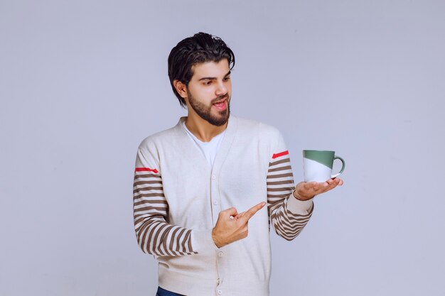 Homem de camisa branca segurando uma xícara de café e apontando para ela.