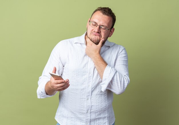 Homem de camisa branca usando óculos segurando um smartphone olhando para ele com expressão cética em pé sobre uma parede verde