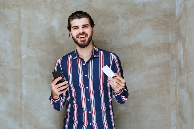 Homem de camisa listrada segurando um cartão de visita e seu smartphone enquanto sorri