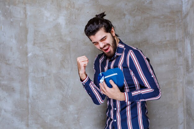 Homem de camisa listrada segurando uma caixa de presente em formato de coração azul e se sentindo bem-sucedido