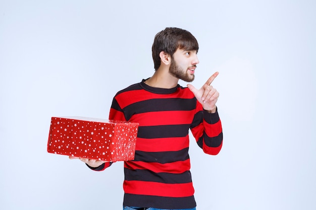 Foto grátis homem de camisa listrada vermelha com uma caixa de presente vermelha e chamando alguém para apresentá-la.