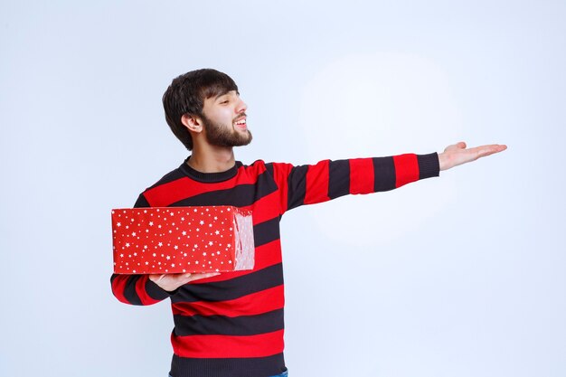 Homem de camisa listrada vermelha com uma caixa de presente vermelha e chamando alguém para apresentá-la.