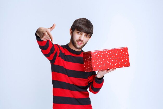 Homem de camisa listrada vermelha com uma caixa de presente vermelha e chamando alguém para apresentá-la.