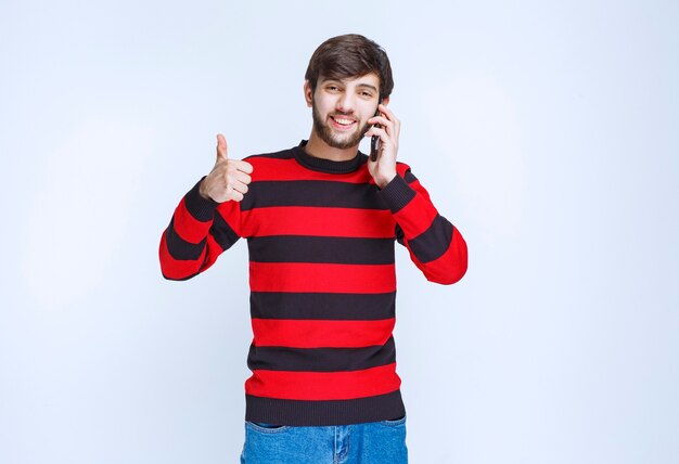 Homem de camisa listrada vermelha, falando ao telefone e mostrando o polegar para ouvir boas notícias.