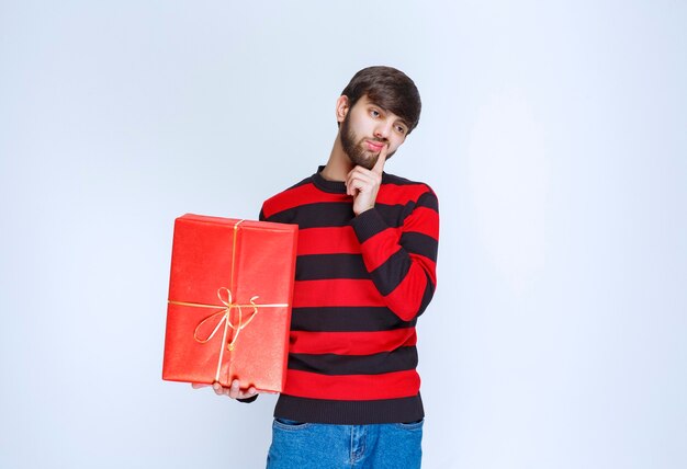 Homem de camisa listrada vermelha segurando uma caixa de presente vermelha e parece confuso e pensativo.